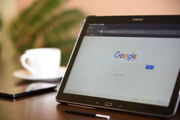 This is image of a samsung tablet on a desk with a cup of coffee displaying the google search engine.