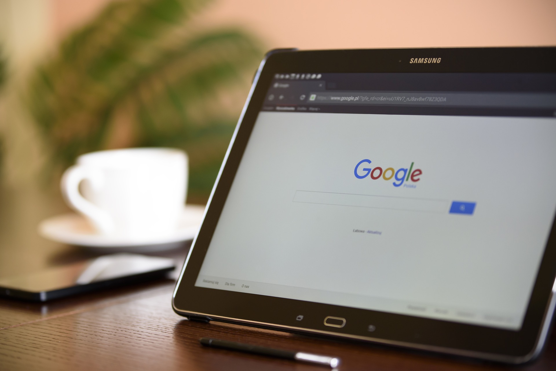 THIS IS IMAGE OF A SAMSUNG TABLET ON A DESK WITH A CUP OF COFFEE DISPLAYING THE GOOGLE SEARCH ENGINE.